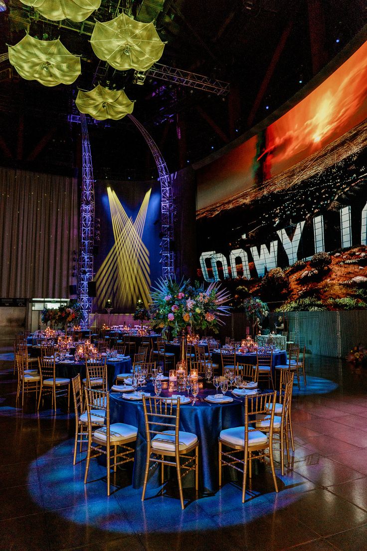 an indoor event venue set up with round tables and blue linens, tall chandeliers and large screen