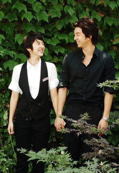 two young men standing next to each other in front of green plants and bushes, one smiling at the camera