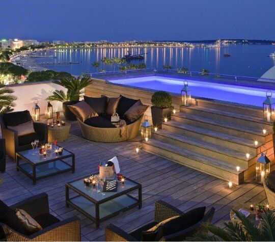 an outdoor deck with lights and seating area next to the water at night, lit up by candles