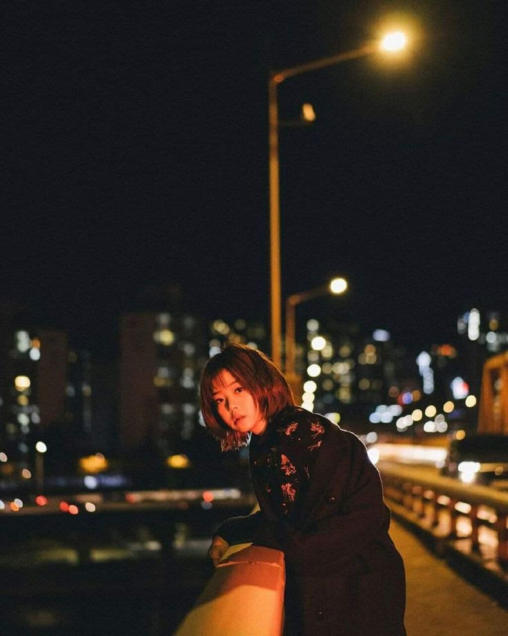 a woman standing on a bridge at night with her hand on the railing and lights in the background