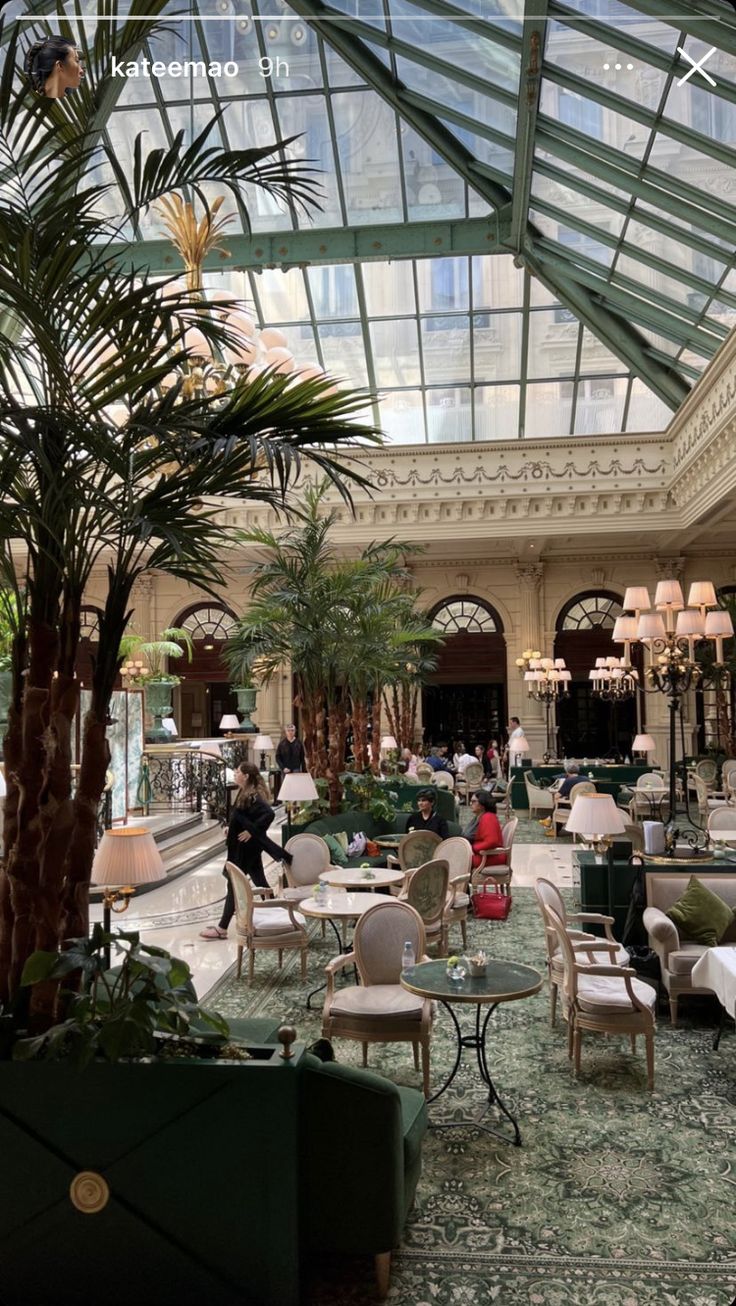 the inside of a hotel lobby with lots of tables and chairs, palm trees and chandeliers