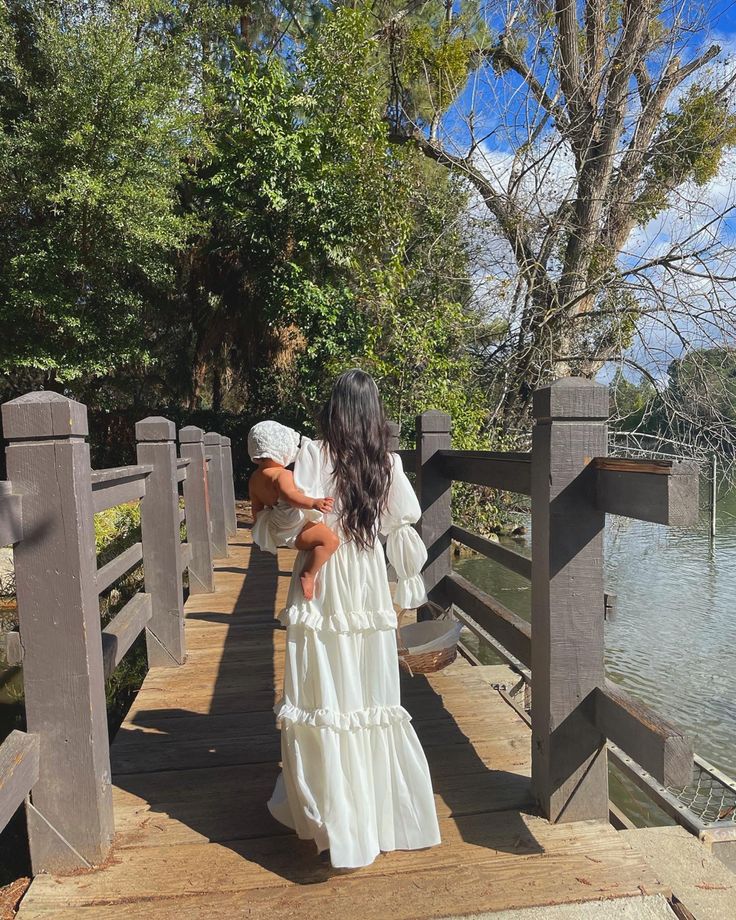 a woman in a white dress is walking on a wooden bridge near the water and trees