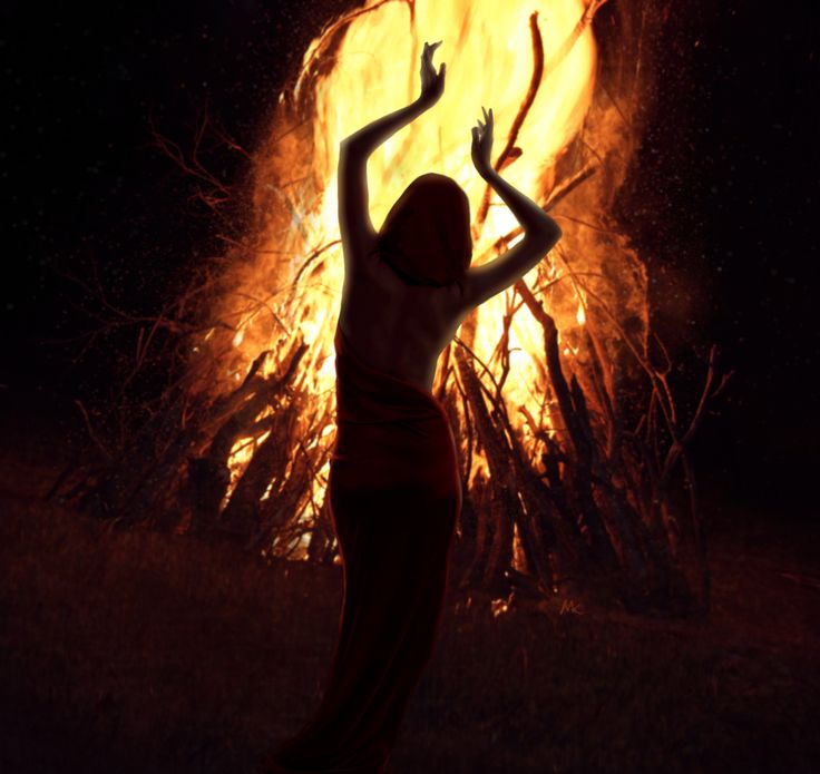 a woman standing in front of a fire with her arms up and hands raised above her head