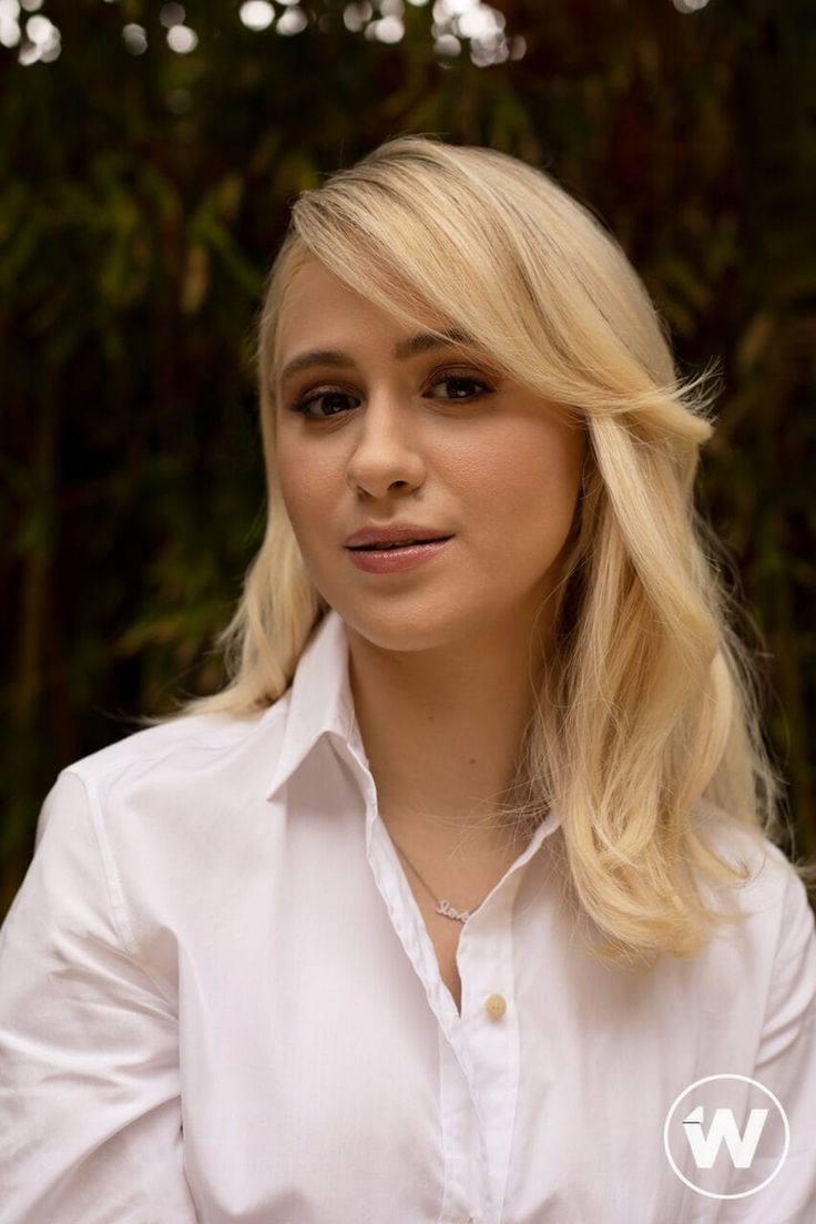 a woman with blonde hair wearing a white shirt and looking at the camera while standing in front of some trees