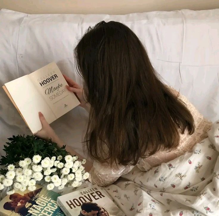 a woman reading a book in bed with flowers and books on the floor next to her