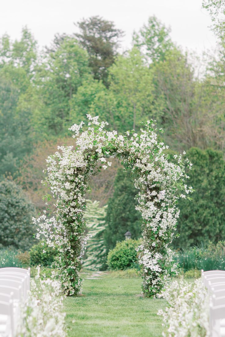 an outdoor ceremony with white chairs and flowers on the arch, in front of trees