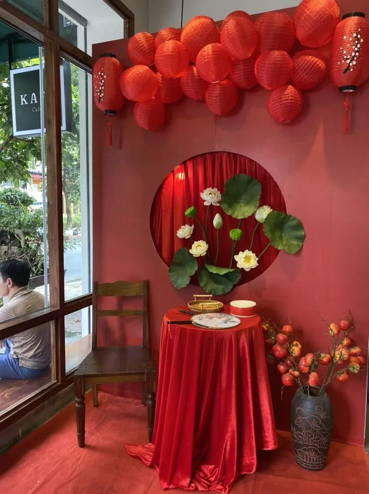 a red table topped with a vase filled with flowers next to a mirror covered in paper lanterns