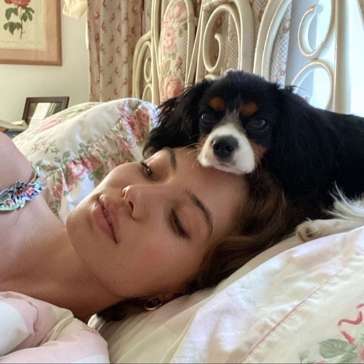 a woman laying in bed with a dog on her head and looking at the camera