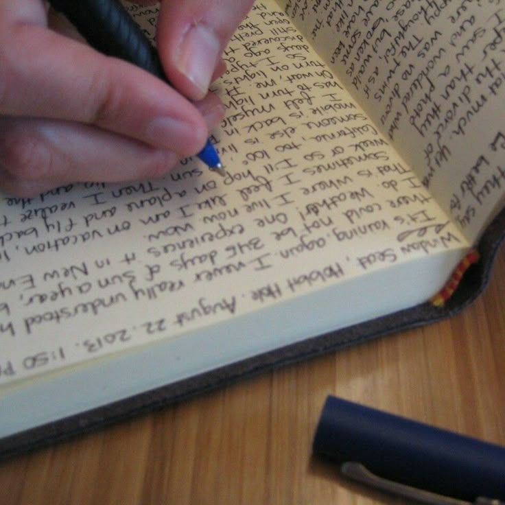 a person writing on an open book with a pen in their left hand, and a small black object next to it