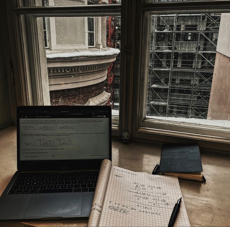 an open notebook sitting on top of a table next to a laptop computer and a window