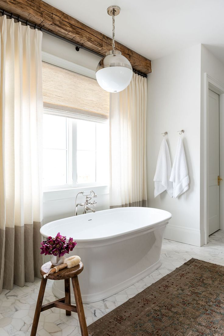 a white bath tub sitting under a window next to a wooden stool with flowers on it