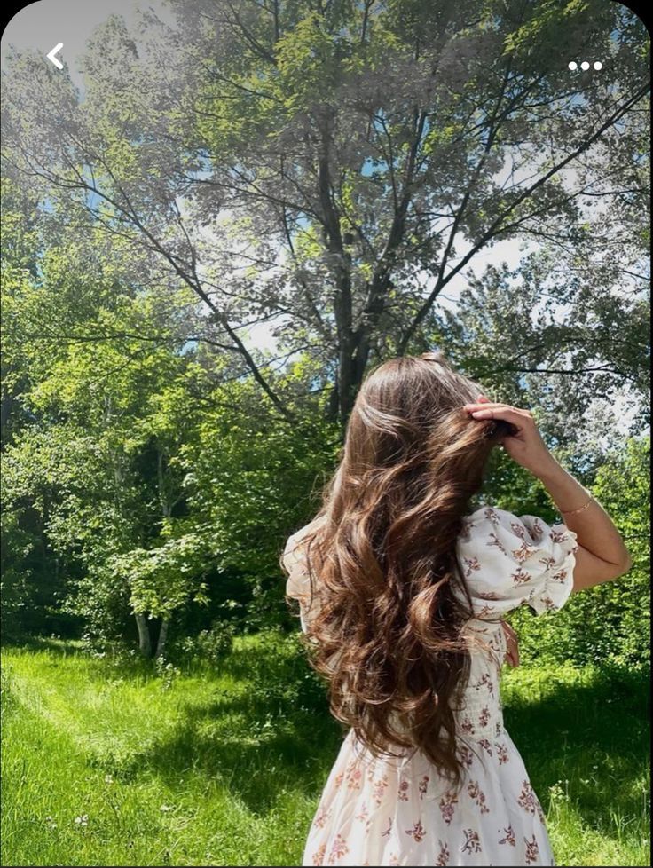 a girl with long hair standing in the grass looking up at a tree and holding her hands to her head