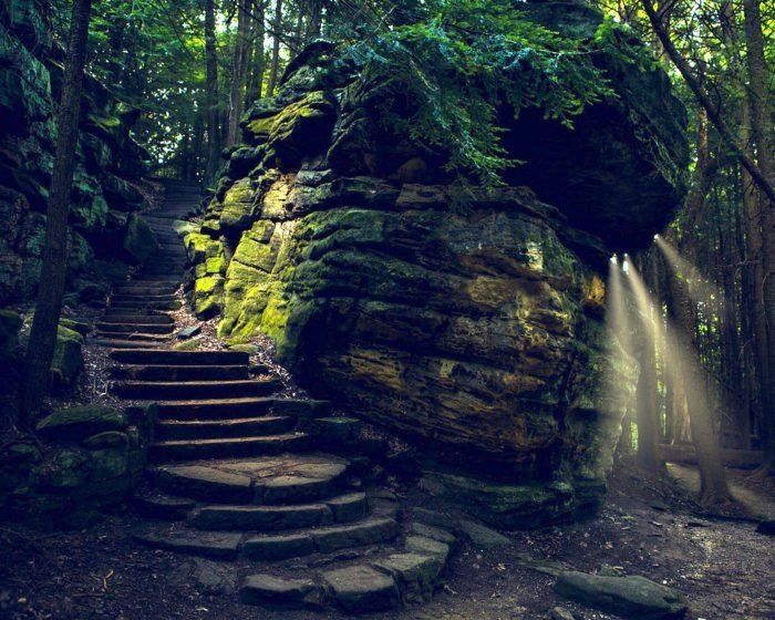 some stairs going up to a big rock in the woods with light coming from it