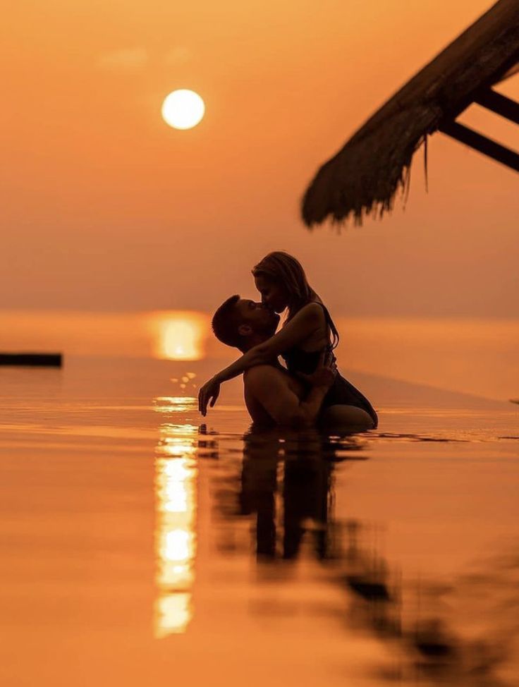 two people are sitting in the water at sunset with their arms around each other and kissing