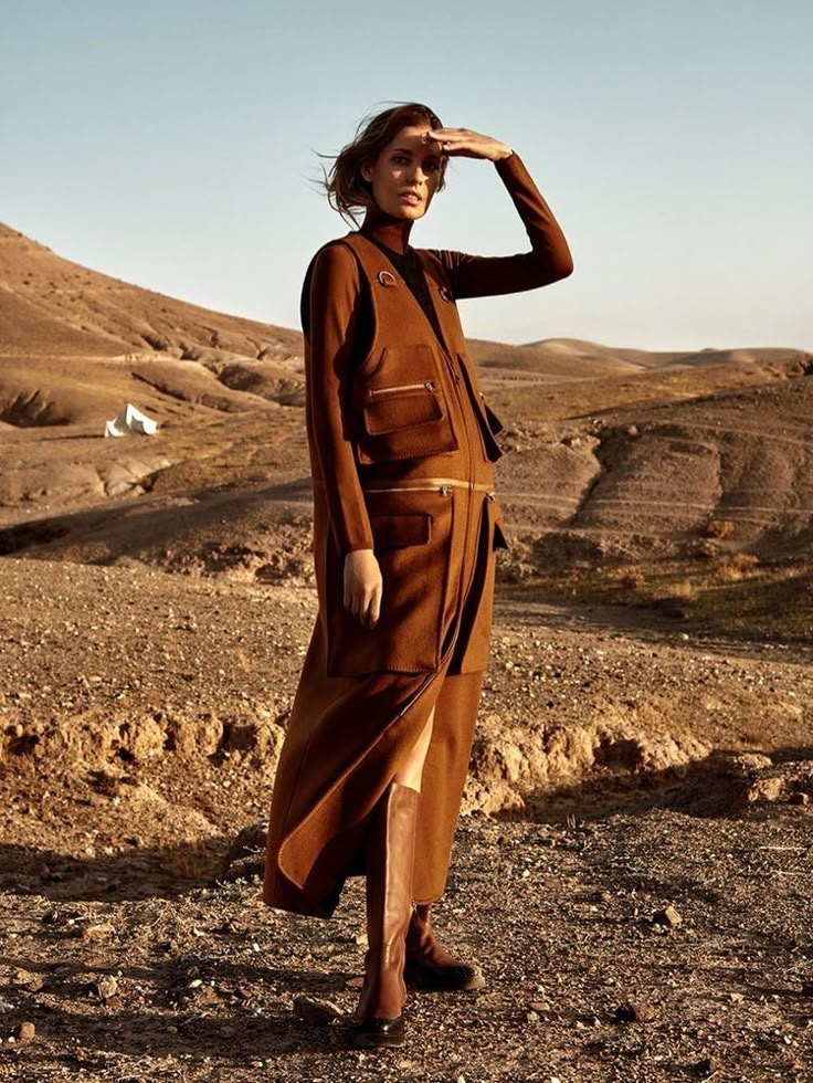 a woman is standing in the desert wearing a brown dress and black boots with her hand on her head