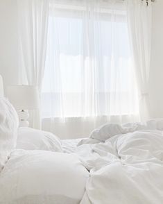 an unmade bed with white linens in front of a window and sheer curtains