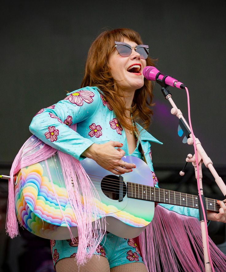 a woman singing into a microphone while holding an acoustic guitar and wearing colorful fringe skirt