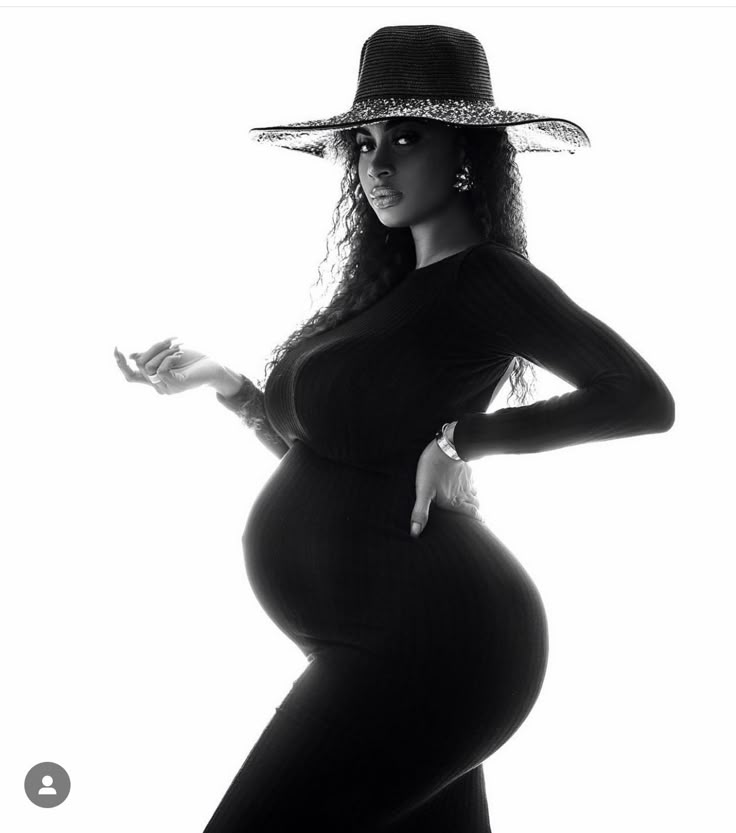 a pregnant woman in a hat poses for a black and white photo with her hands on her hips