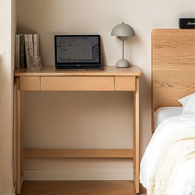 a laptop computer sitting on top of a wooden desk next to a bed with white sheets