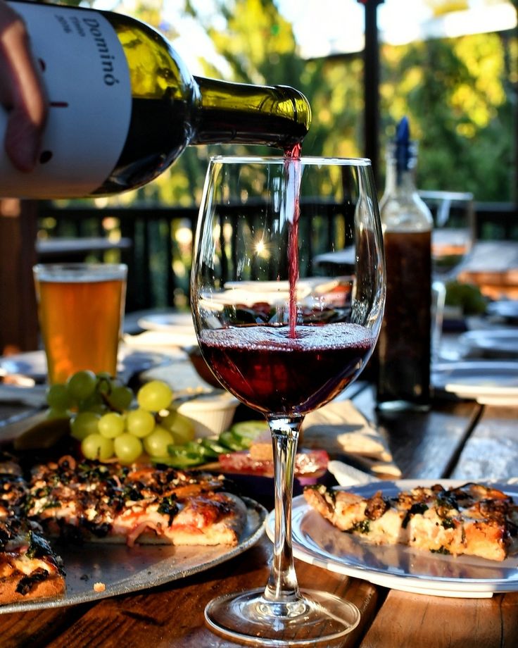 a person pouring wine into a glass on top of a table with pizza and grapes