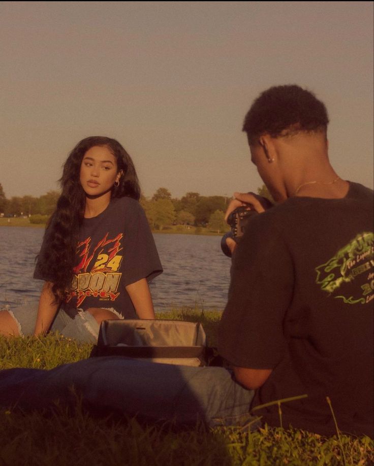 two people sitting on the grass near water and one person taking a photo with a camera