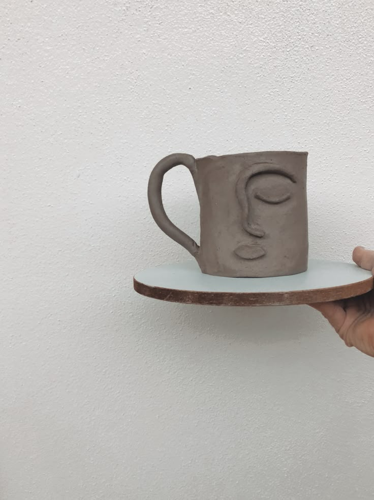 a hand holding a gray coffee cup on top of a white plate against a wall