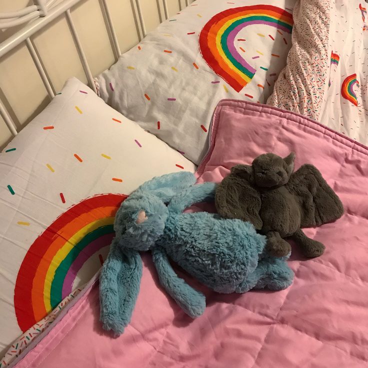 two stuffed animals are laying on a pink bed with white sheets and rainbows in the background