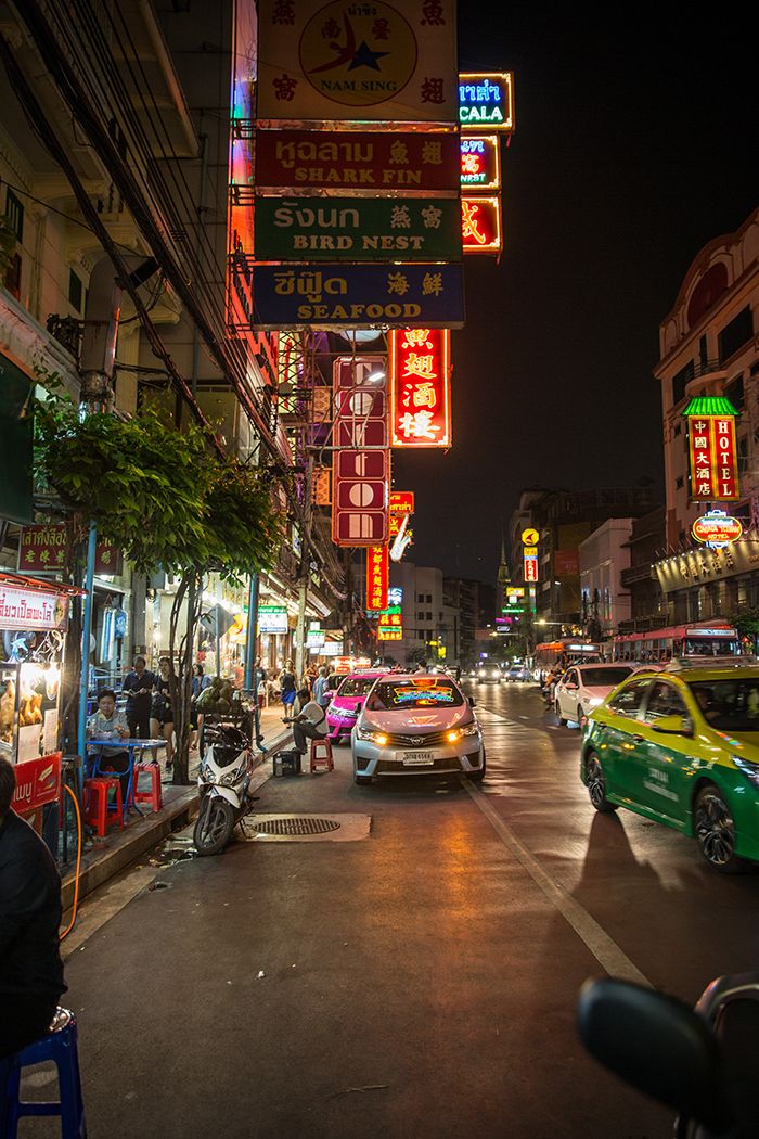 cars driving down the street at night with neon signs on buildings and people sitting in chairs