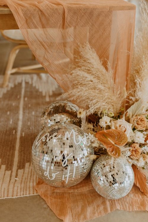 two disco ball ornaments sitting on top of a wooden table next to flowers and feathers