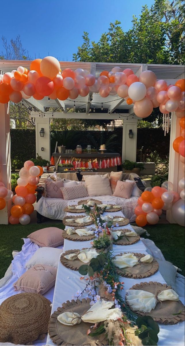 an outdoor table set up for a party with orange and white balloons on the ceiling