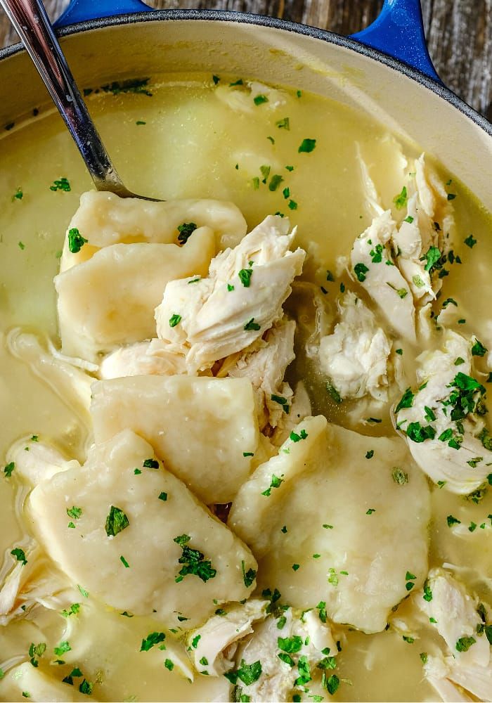 a blue pot filled with chicken and dumplings on top of a wooden table next to a spoon