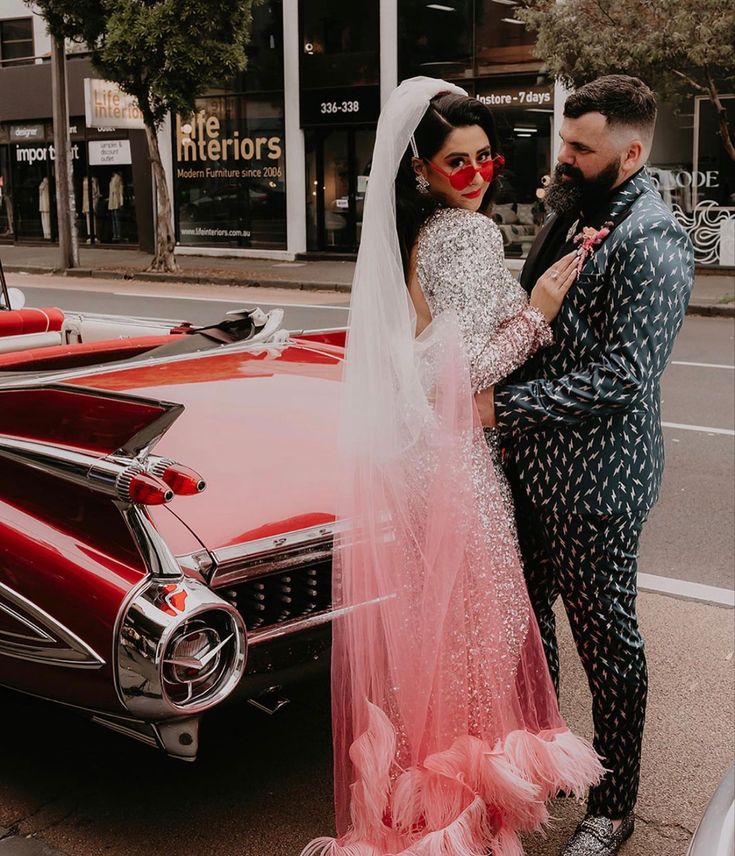 a bride and groom standing in front of an old red car on the side of the road