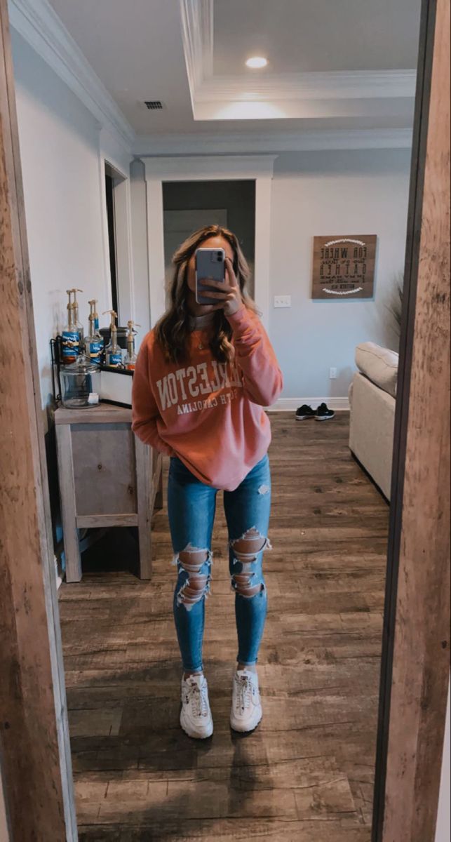 a woman taking a selfie in the mirror wearing ripped jeans and an orange sweatshirt