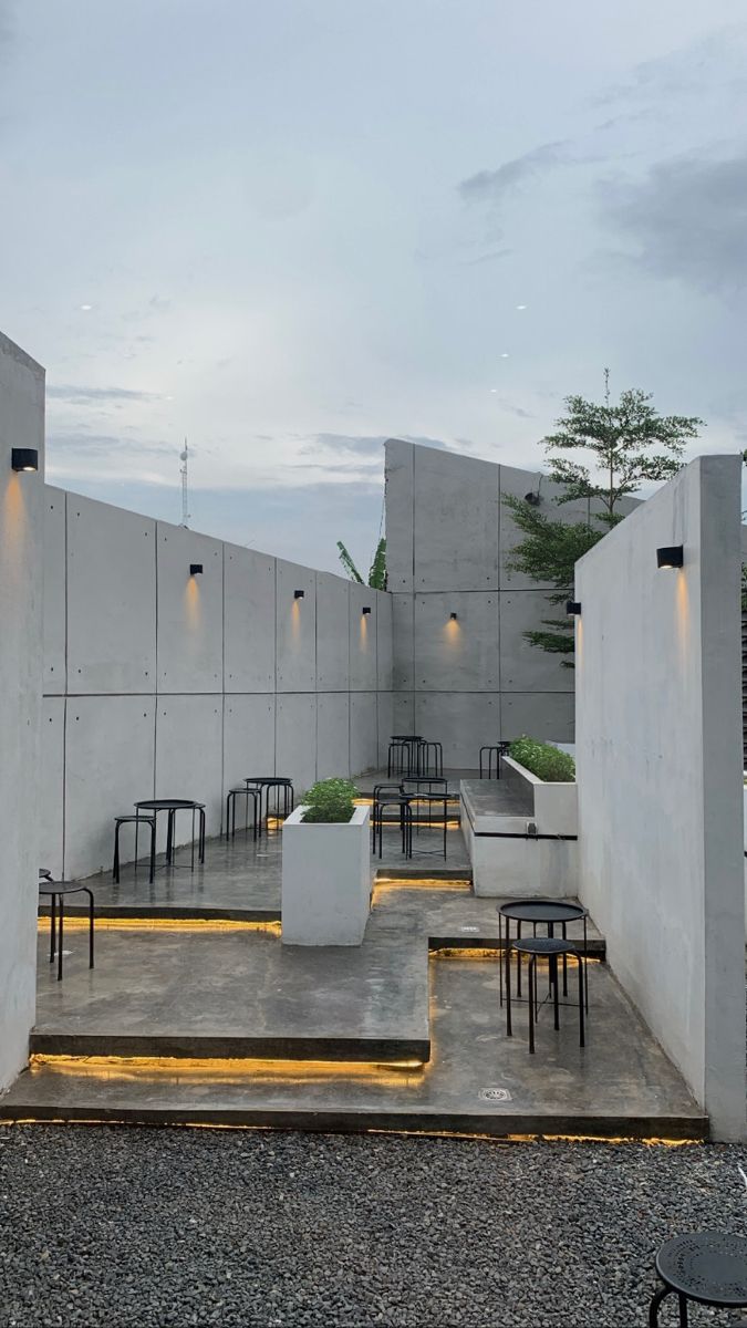 an outdoor seating area with tables and chairs in front of a concrete wall at dusk