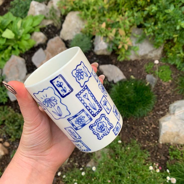 a hand holding a blue and white coffee cup in front of some rocks, grass and flowers