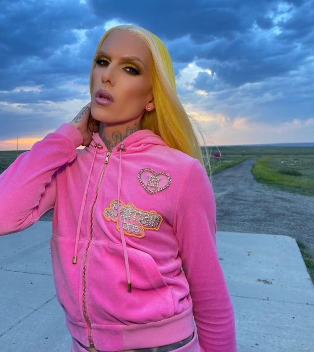 a woman with blonde hair wearing a pink hoodie and silver pants standing in front of a dirt road