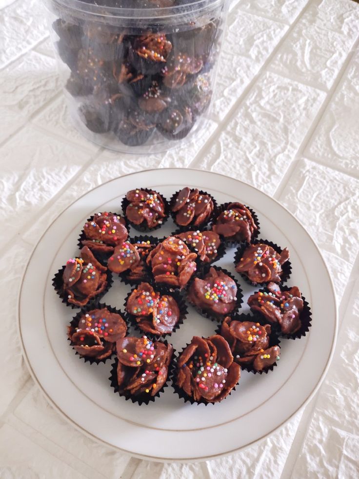 a plate full of chocolate covered cupcakes next to a plastic container filled with sprinkles