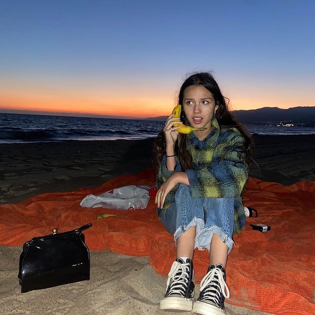 a woman is sitting on the beach and holding a banana up to her ear as she looks at the camera