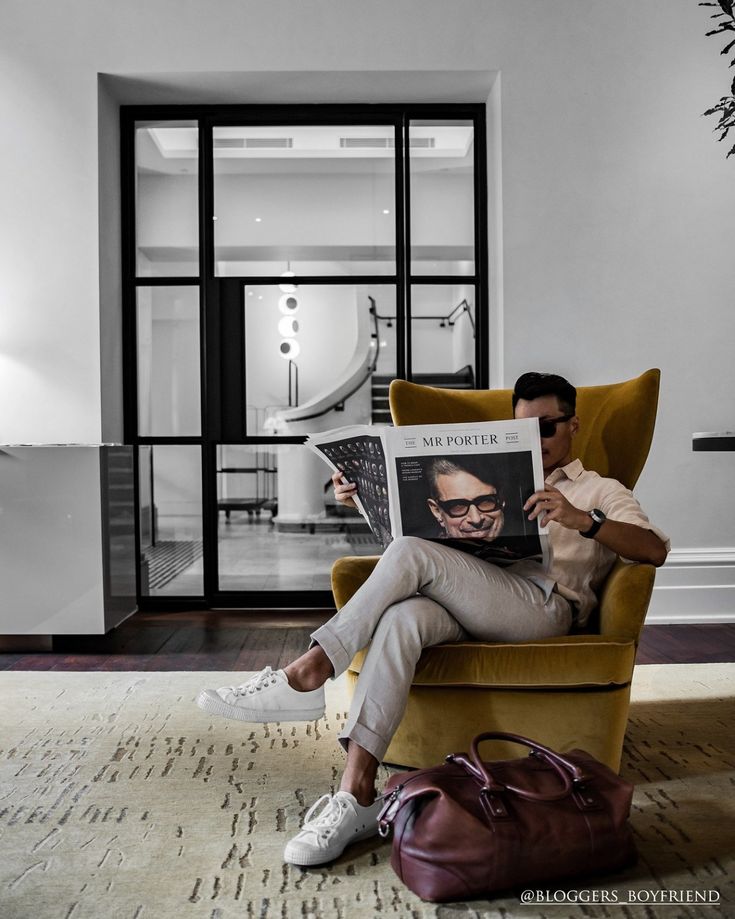 a man sitting in a chair with a magazine on his lap and a handbag next to him