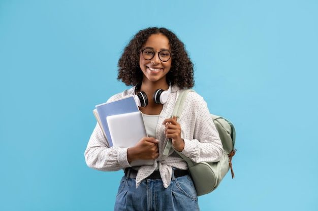 a smiling woman with headphones and books in her hands is standing against a blue background