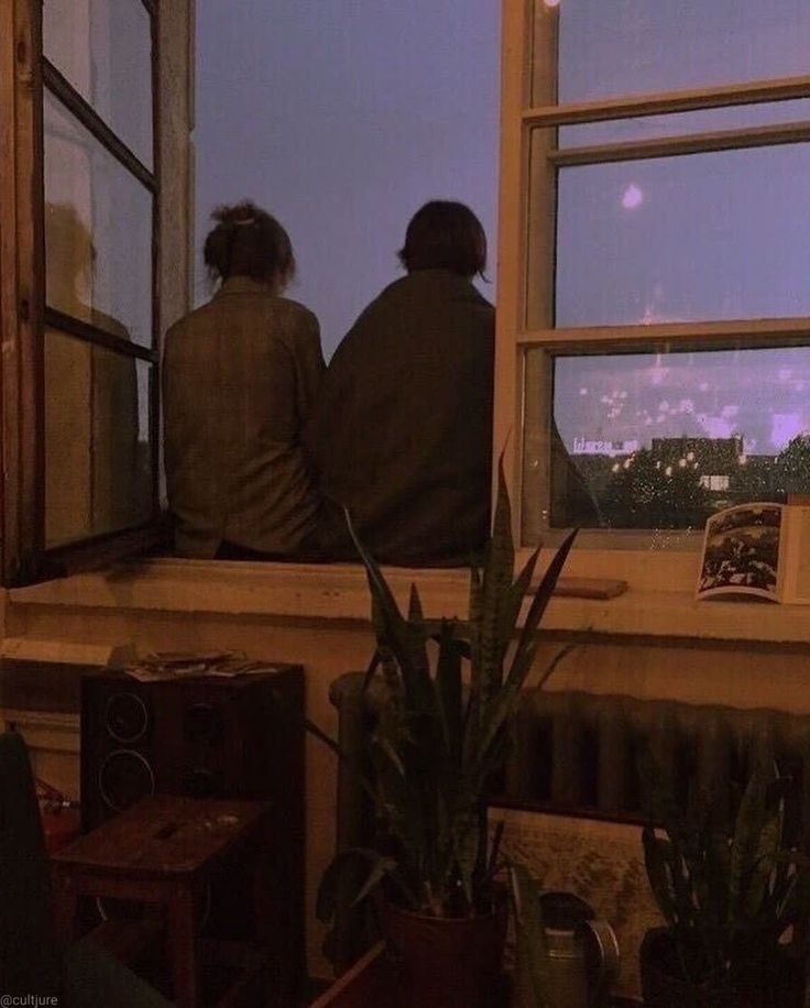 two people are sitting on a window sill looking out at the city below them