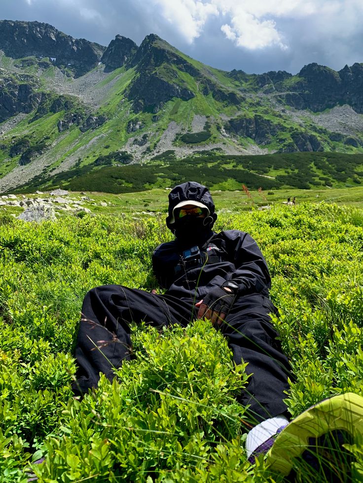 a man sitting on top of a lush green field next to a tall mountain covered in clouds