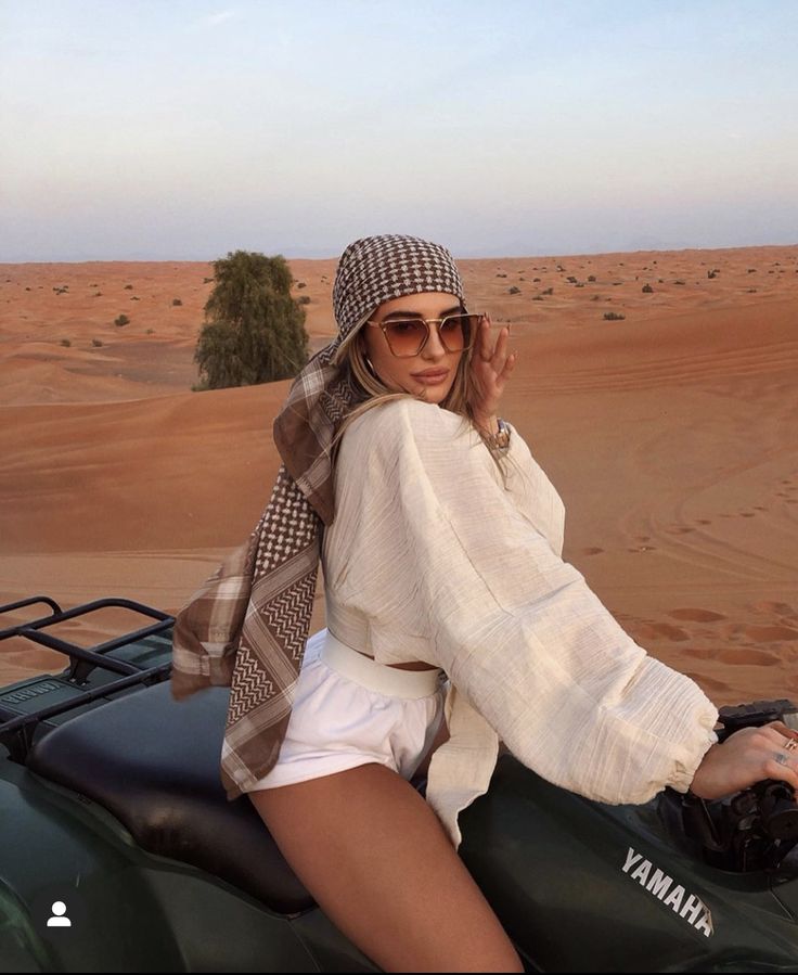 a woman sitting on top of a green jeep in the desert wearing sunglasses and a scarf