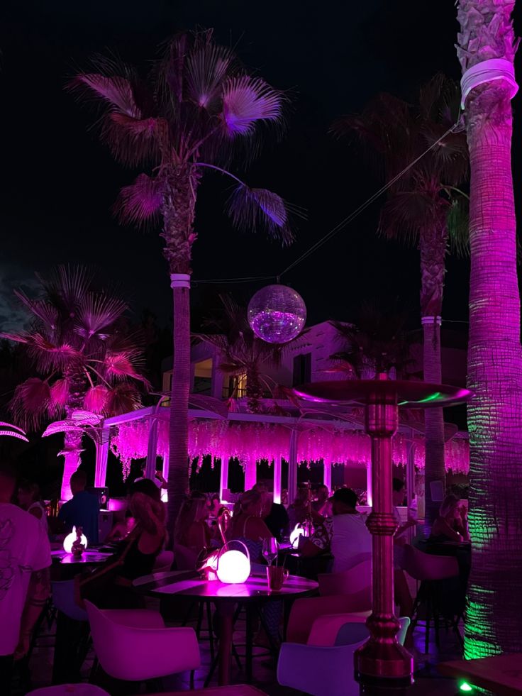 people sitting at tables under palm trees with purple lighting in the dark sky behind them