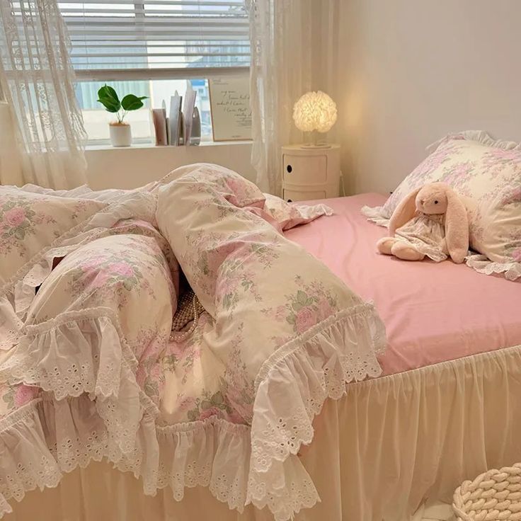 a pink bed with white ruffles and lace on the bedspread is in front of a window