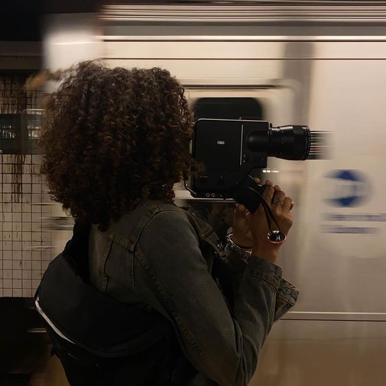 a woman with curly hair is holding a camera in front of a moving subway train