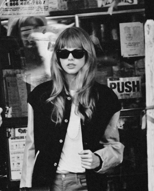 black and white photo of a woman in sunglasses standing on the sidewalk with her hands in her pockets