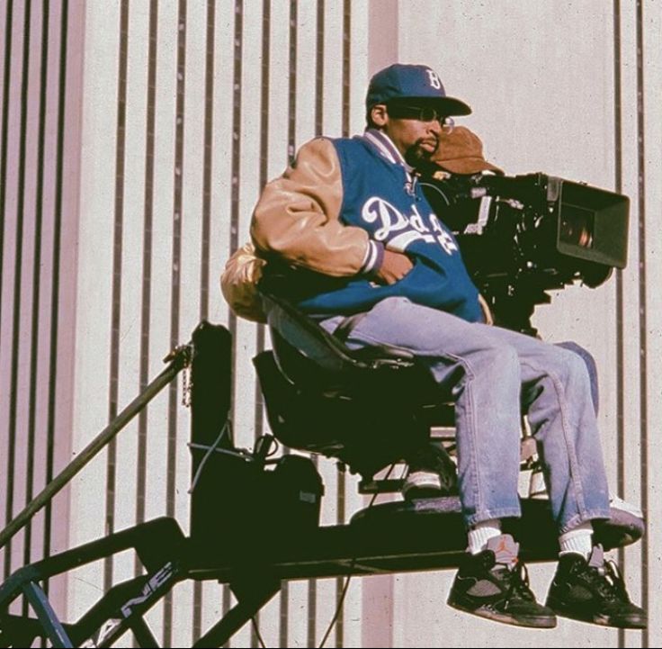 a man sitting in a chair with a camera on his shoulder and wearing a baseball cap