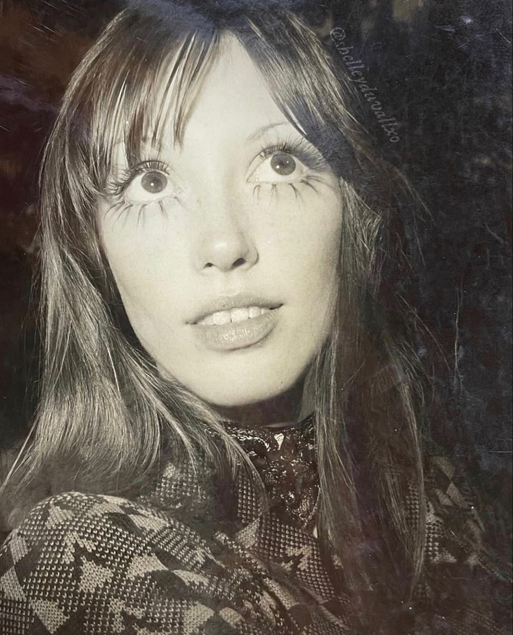 a black and white photo of a woman with long hair wearing a patterned shirt looking at the camera