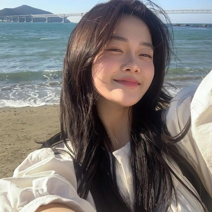 a woman with long black hair standing on the beach next to the ocean and looking at the camera